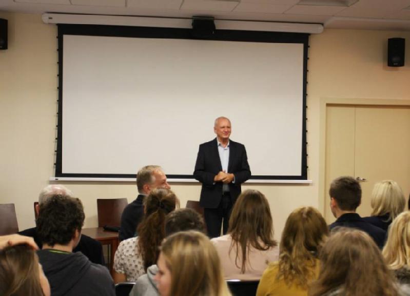 Mężczyzna w garniturze stoi i uśmiecha się przed pustym ekranem projektora, zwracając się do siedzącej publiczności w klasie lub sali seminaryjnej.