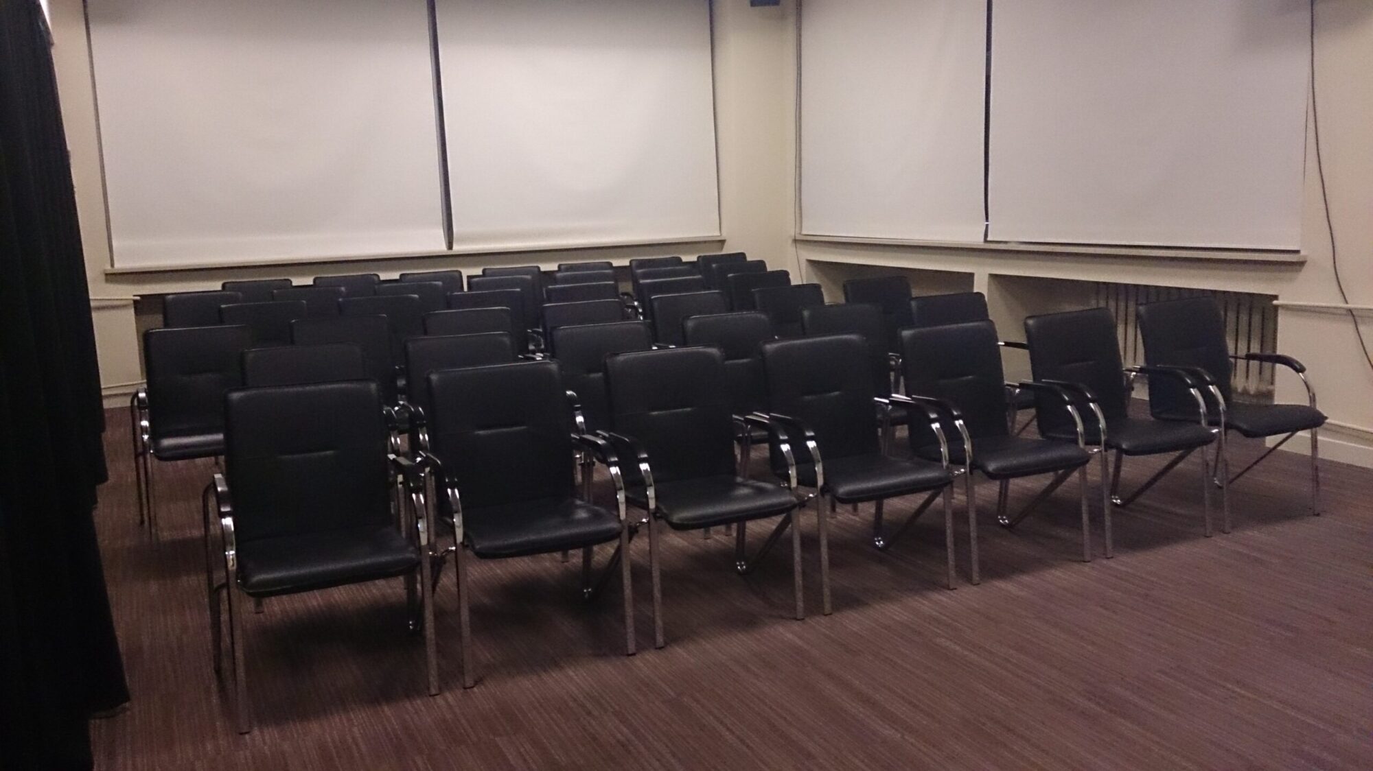 W małej sali konferencyjnej ustawione są rzędy pustych, czarnych krzeseł. Okna są zasłonięte białymi żaluzjami, a podłoga wyłożona jest brązowym dywanem.
