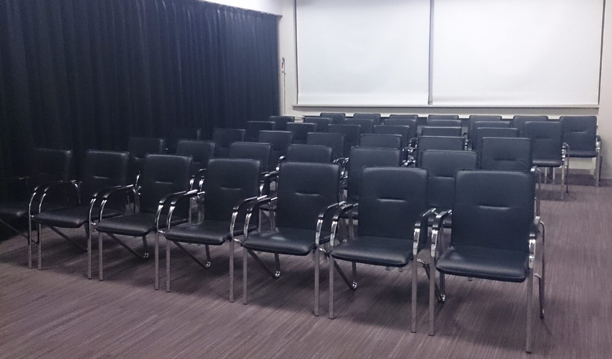 Rzędy pustych, czarnych krzeseł z metalowymi podłokietnikami stoją starannie ustawione w pokoju z brązowym dywanem, ciemnymi zasłonami po jednej stronie i pustym, białym ekranem projekcyjnym z przodu.