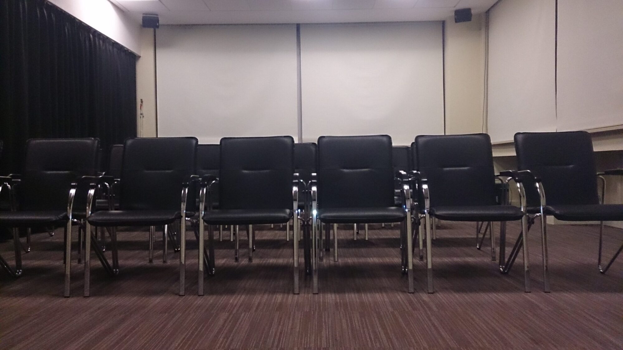 W małej sali konferencyjnej lub sali spotkań ustawione są rzędy pustych czarnych krzeseł z metalowymi podłokietnikami, z zasłoniętymi żaluzjami i wykładziną dywanową.