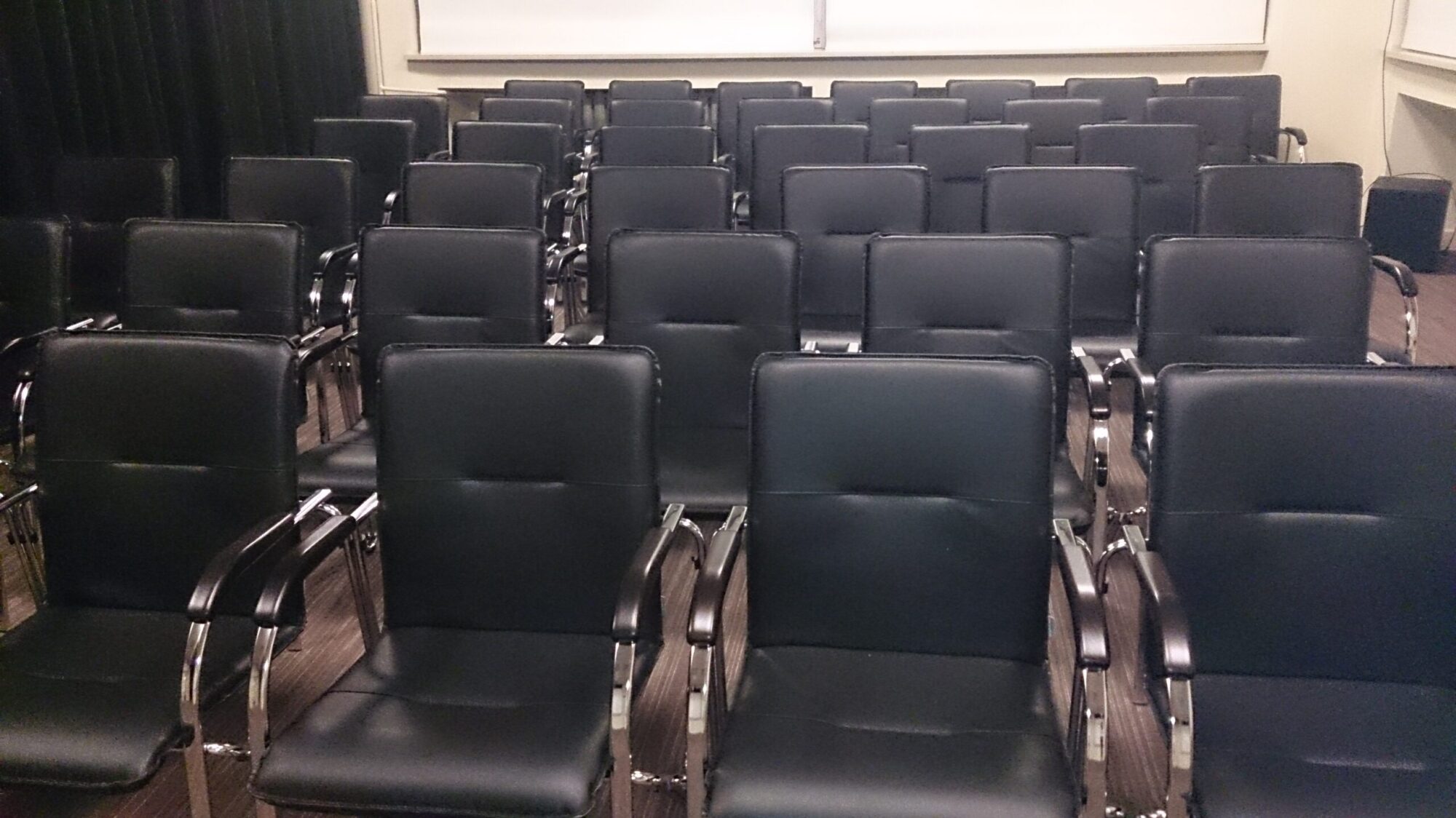 W małej sali konferencyjnej lub sali spotkań ustawione są rzędy pustych, czarnych skórzanych krzeseł z metalowymi podłokietnikami, z przodu znajduje się tablica lub ekran.
