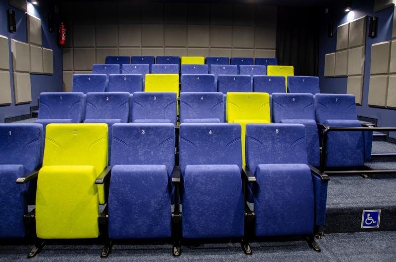 Niewielkie kino z rzędami niebieskich siedzeń i kilkoma jaskrawożółtymi siedzeniami; w pierwszym rzędzie widoczna jest dostępna strefa z miejscem dla osób na wózkach inwalidzkich.