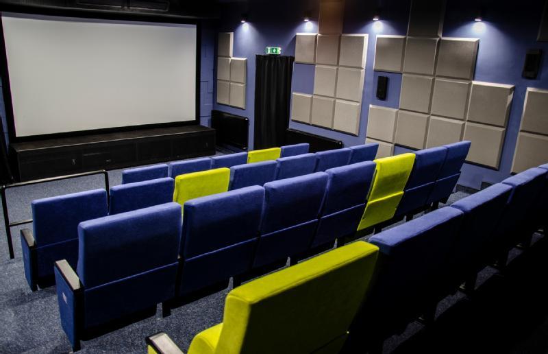 Małe nowoczesne kino z rzędami niebieskich siedzeń, z których dwa są jasnożółte, zwrócone w stronę dużego, pustego ekranu. Ściany pokryte są szarymi panelami akustycznymi, a górne światła delikatnie oświetlają pomieszczenie.
