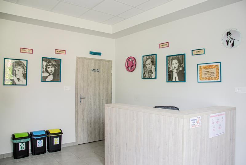 Nowoczesna recepcja z jasnym drewnianym biurkiem, pojemnikami na odpady do recyklingu, oprawionymi portretami retro na ścianach i zamkniętymi drzwiami z napisem „MANAGER”. Przestrzeń jest czysta, jasna i uporządkowana.