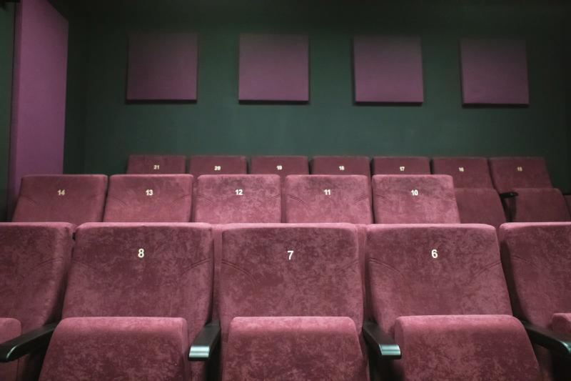 Rzędy pustych bordowych foteli teatralnych z widocznymi numerami miejsc, naprzeciwko ciemnozielonej ściany z panelami dźwiękochłonnymi. Teatr jest pusty i cicho oświetlony.
