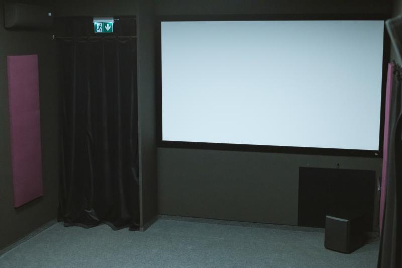 Mały, pusty pokój kina domowego z pustym ekranem projekcyjnym, ciemnymi ścianami, wykładziną dywanową, fioletowymi panelami akustycznymi, czarną zasłoną i pojedynczym głośnikiem na podłodze. Nad zasłoną widoczny jest znak wyjścia.