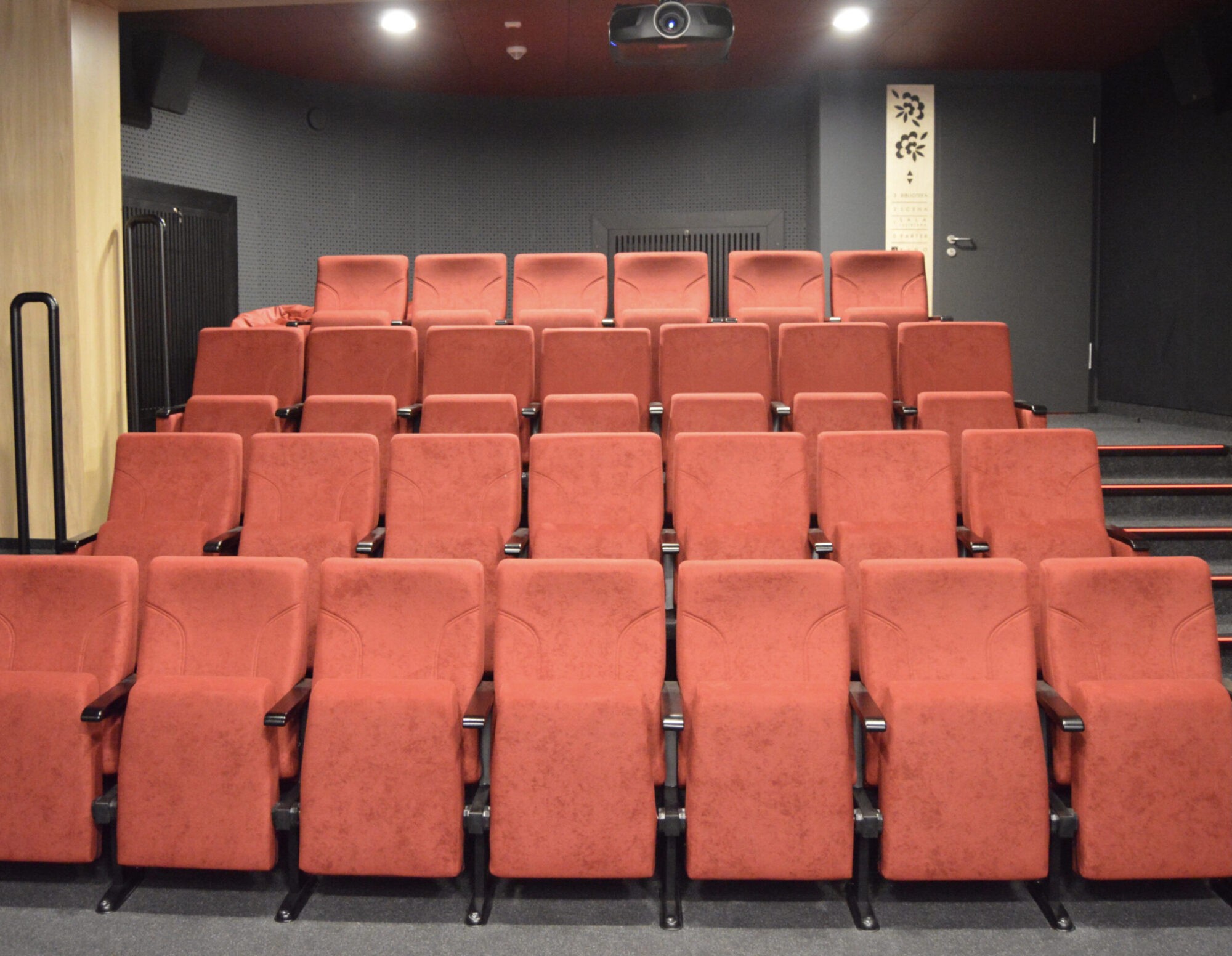 Niewielka sala kinowa z czterema rzędami pustych, czerwonych foteli, projektorem na suficie, schodami po prawej stronie i drzwiami z tyłu, nad którymi wisi ozdobny szyld.