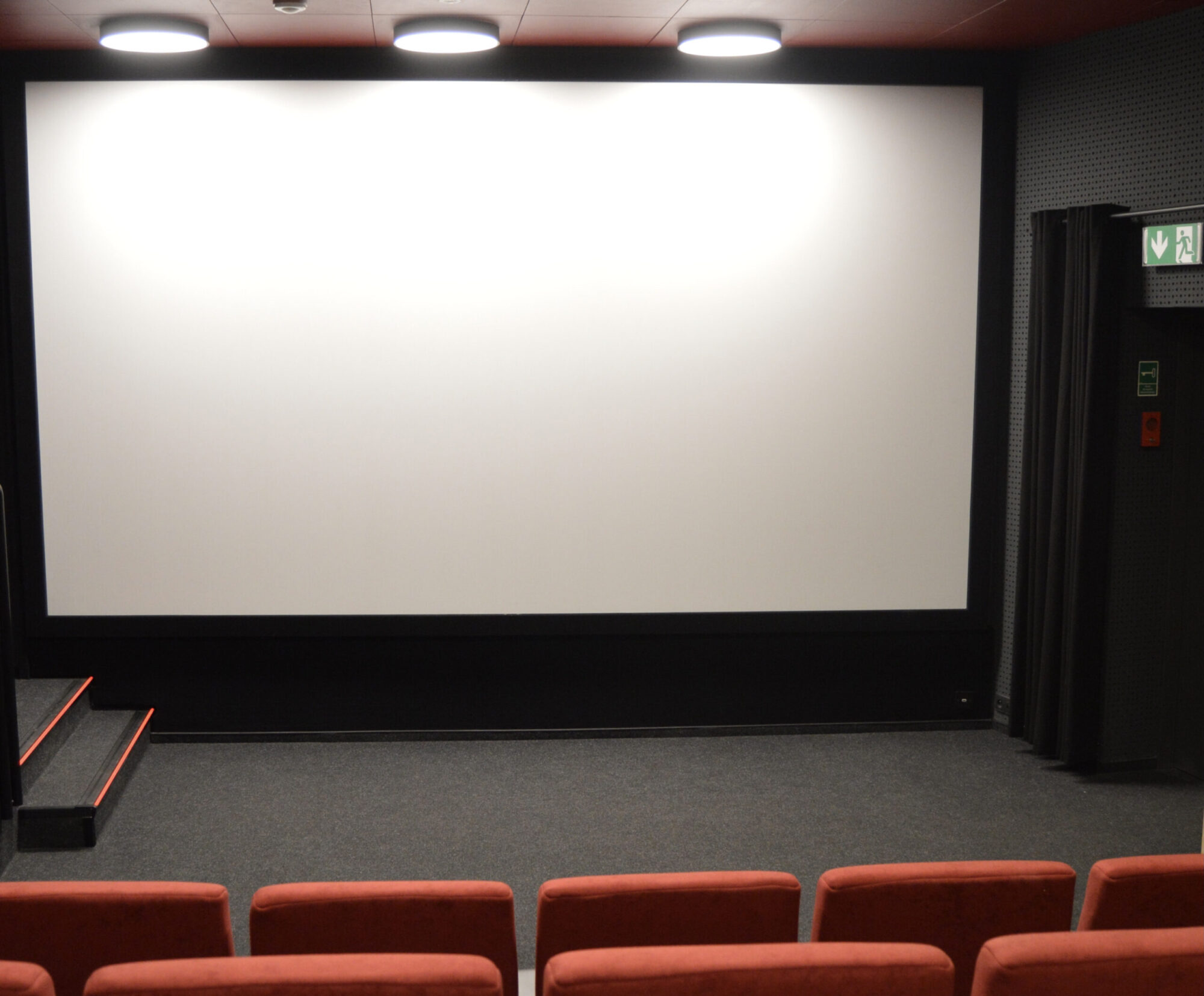Puste kino z czerwonymi siedzeniami zwróconymi w stronę dużego, białego ekranu. Sala jest słabo oświetlona lampami sufitowymi, a znak wyjścia jest widoczny po prawej stronie ekranu.