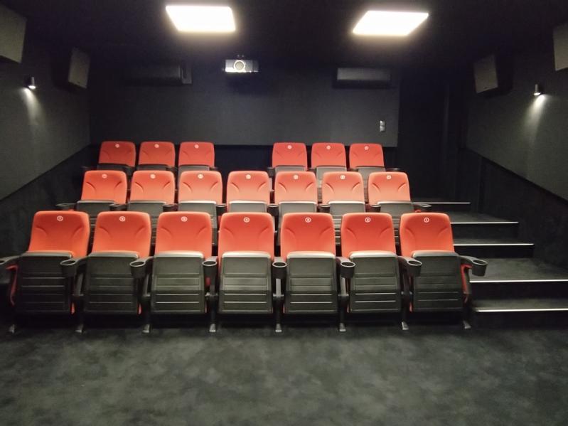 Małe, puste kino z trzema rzędami czerwonych siedzeń, czarnymi podłokietnikami i wykładziną. W pomieszczeniu jest słabe oświetlenie, czarne ściany i projektor zamontowany na tylnej ścianie.
