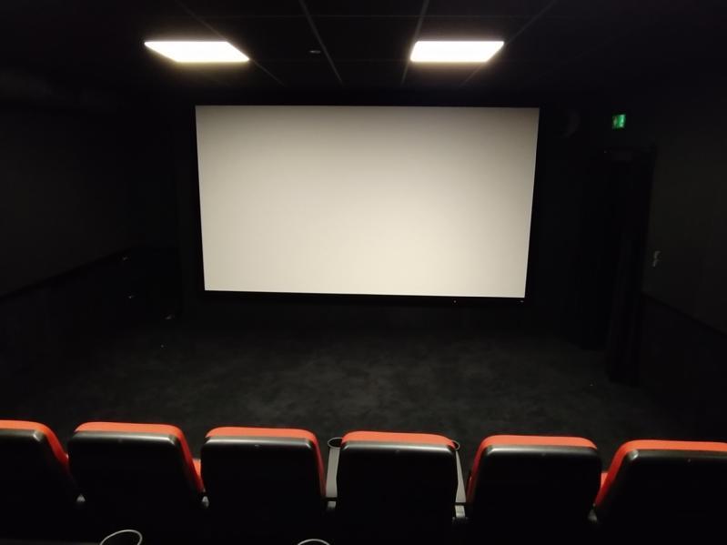 Małe, puste kino z pięcioma czerwonymi i czarnymi siedzeniami naprzeciwko dużego, pustego ekranu pod oświetleniem sufitowym i ciemnymi ścianami tworzącymi przytulną atmosferę.