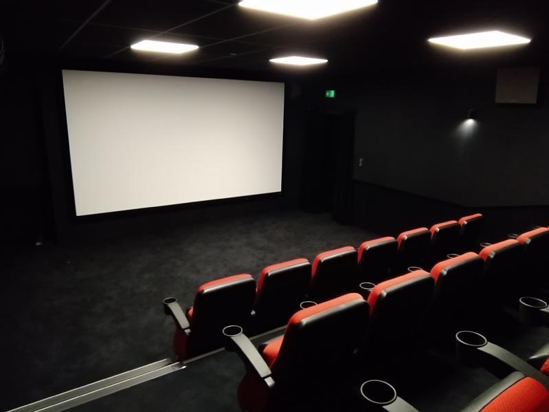 Małe, puste kino z czerwonymi i czarnymi siedzeniami zwróconymi w stronę dużego białego ekranu. Pomieszczenie jest słabo oświetlone, a ściany i podłoga są ciemne, co tworzy przytulną atmosferę.