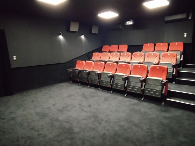 Małe, nowoczesne kino z dwoma rzędami czerwonych siedzeń na czarnej wykładzinie, przyćmionym oświetleniem i ciemnym wnętrzem. Sala wydaje się pusta i cicha.