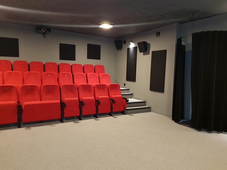 Mała, nowoczesna sala kinowa z rzędami czerwonych foteli teatralnych, szarymi ścianami, panelami akustycznymi i czarną zasłoną po prawej stronie. Sala jest pusta i dobrze oświetlona.