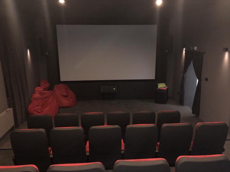 Małe, słabo oświetlone kino domowe z rzędami czarnych siedzeń z czerwonymi wykończeniami zwróconych w stronę dużego, pustego ekranu. W przednim rogu ustawiono czerwone worki z fasolą, a przy ciemnych drzwiach znajduje się mały stolik z poduszkami.