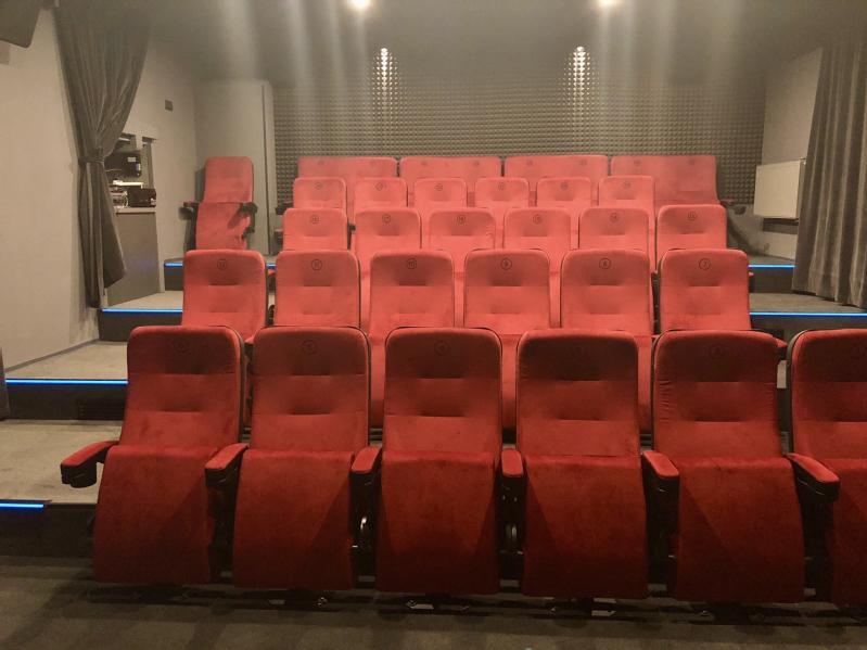 Puste małe kino z rzędami czerwonych siedzeń, słabym oświetleniem, szarymi ścianami i zasłonami po bokach. Niebieskie światła oświetlają stopnie między rzędami siedzeń.