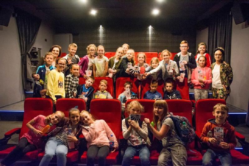 Grupa dzieci i kilku dorosłych pozuje do zdjęcia, uśmiechając się i trzymając przedmioty, siedząc lub stojąc w małym kinie z czerwonymi siedzeniami i ciemnymi ścianami.