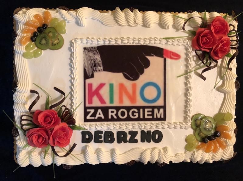 Prostokątny tort ozdobiony lukrem w kształcie czerwonej róży, plasterkami owoców i białym lukrem. Na środku znajduje się logo KINO ZA ROGIEM i wskazująca dłoń, a poniżej napis DEBRZNO.