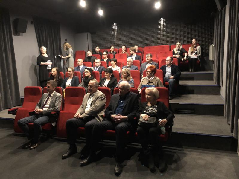 Grupa osób siedzi na czerwonych siedzeniach w małym teatrze lub kinie, zwrócona twarzą do przodu; niektórzy rozmawiają, podczas gdy kilku stoi z tyłu przy wejściu.