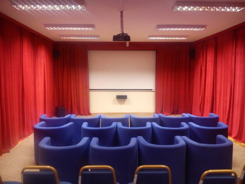 Niewielka sala kinowa z rzędami niebieskich, wyściełanych foteli zwróconych w stronę białego ekranu projekcyjnego, otoczona czerwonymi zasłonami i oświetlona lampami sufitowymi.