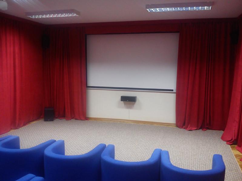 Niewielkie kino domowe z niebieskimi, wyściełanymi siedzeniami naprzeciwko pustego ekranu projekcyjnego, otoczone czerwonymi zasłonami i neutralną wykładziną dywanową.
