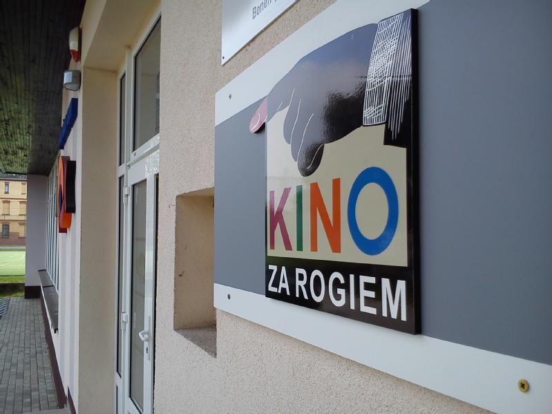 Znak na ścianie budynku pokazuje rękę wskazującą na tekst, który kolorowymi literami głosi Kino za Rogiem. W tle widać szklane drzwi i kolejny znak.