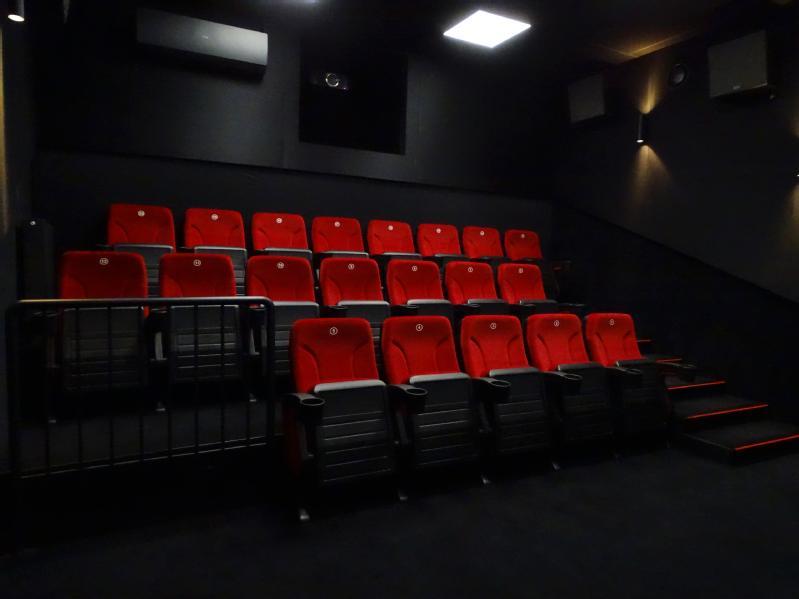 Małe, ciemne kino z trzema rzędami pustych czerwonych siedzeń zwróconych w stronę ekranu. Pomieszczenie jest słabo oświetlone, a ściany i podłoga są czarne. Schody z czerwonymi krawędziami prowadzą do górnych rzędów.