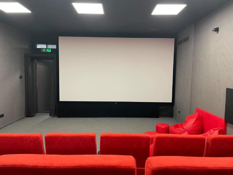 Mała sala kinowa z czerwonymi, wyściełanymi siedzeniami, dużym pustym ekranem z przodu, szarymi ścianami i kwadratowymi lampami sufitowymi. Czerwony fotel-worek ustawiony jest przy ekranie po prawej stronie.