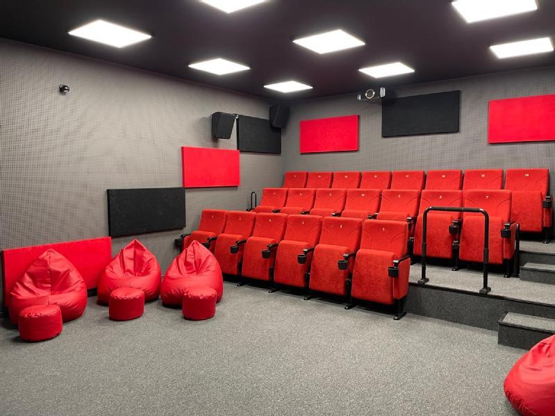 Niewielkie, nowoczesne kino domowe z czerwonymi, wyściełanymi siedzeniami ustawionymi w rzędach, czterema czerwonymi fotelami typu bean bag z przodu, szarymi ścianami z czarnymi i czerwonymi panelami akustycznymi oraz projektorem zamontowanym na suficie.