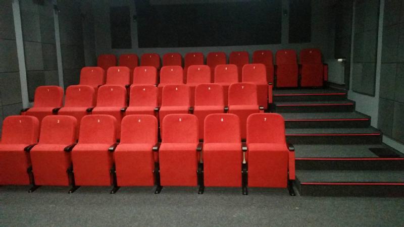 Rzędy pustych, czerwonych foteli teatralnych ustawiono w małej sali widowiskowej z szarymi ścianami i wyłożonymi wykładziną schodami po prawej stronie.