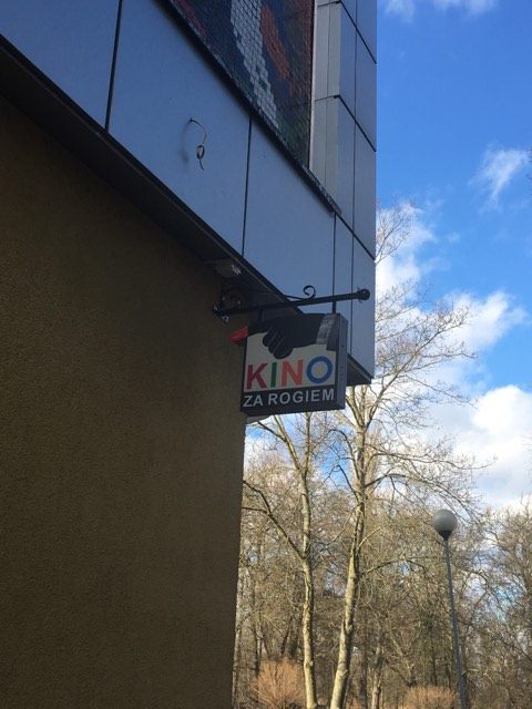 Na ścianie budynku wisi niewielki szyld z napisem Kino Za Rogiem, w tle widać drzewa i latarnię uliczną pod częściowo zachmurzonym niebem.