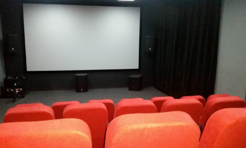 Rzędy pustych, czerwonych siedzeń w teatrze ustawione są naprzeciwko dużego, pustego ekranu projekcyjnego w małej, słabo oświetlonej sali kinowej z czarnymi ścianami, głośnikami i dwoma subwooferami w pobliżu ekranu.