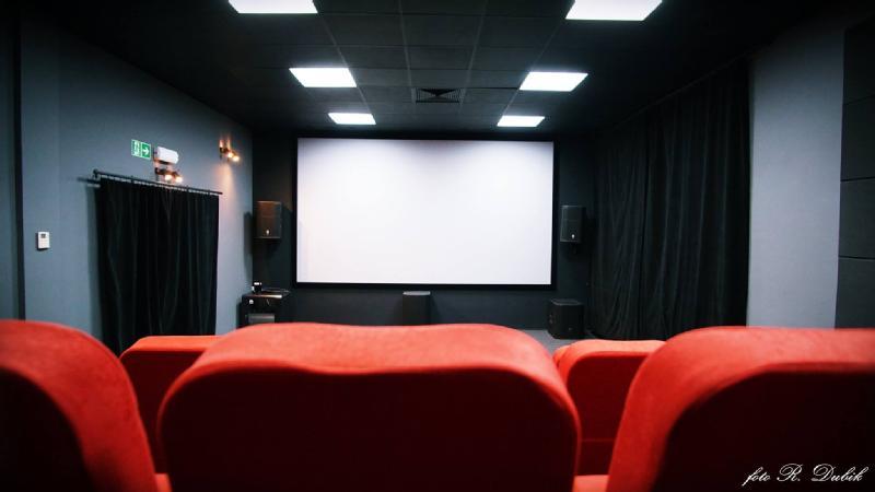 Małe kino z czerwonymi siedzeniami zwróconymi w stronę dużego, pustego ekranu, otoczone ciemnymi ścianami i sufitowymi lampami. Na bocznych ścianach znajdują się głośniki, a przy wyjściu czarna zasłona.