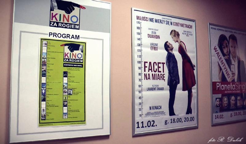 Plakaty na ścianie reklamują filmy w kinie. Lewy plakat zawiera listę programów kinowych; środkowy przedstawia parę stojącą naprzeciwko siebie; prawe plakaty wyświetlają tytuły filmów i wizerunki aktorów.