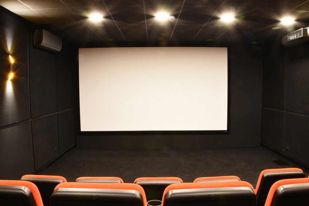 Niewielkie kino z czerwonymi i czarnymi, wyściełanymi siedzeniami naprzeciwko dużego, pustego ekranu projekcyjnego, ciemnymi ścianami i oświetleniem sufitowym oświetlającym pomieszczenie.
