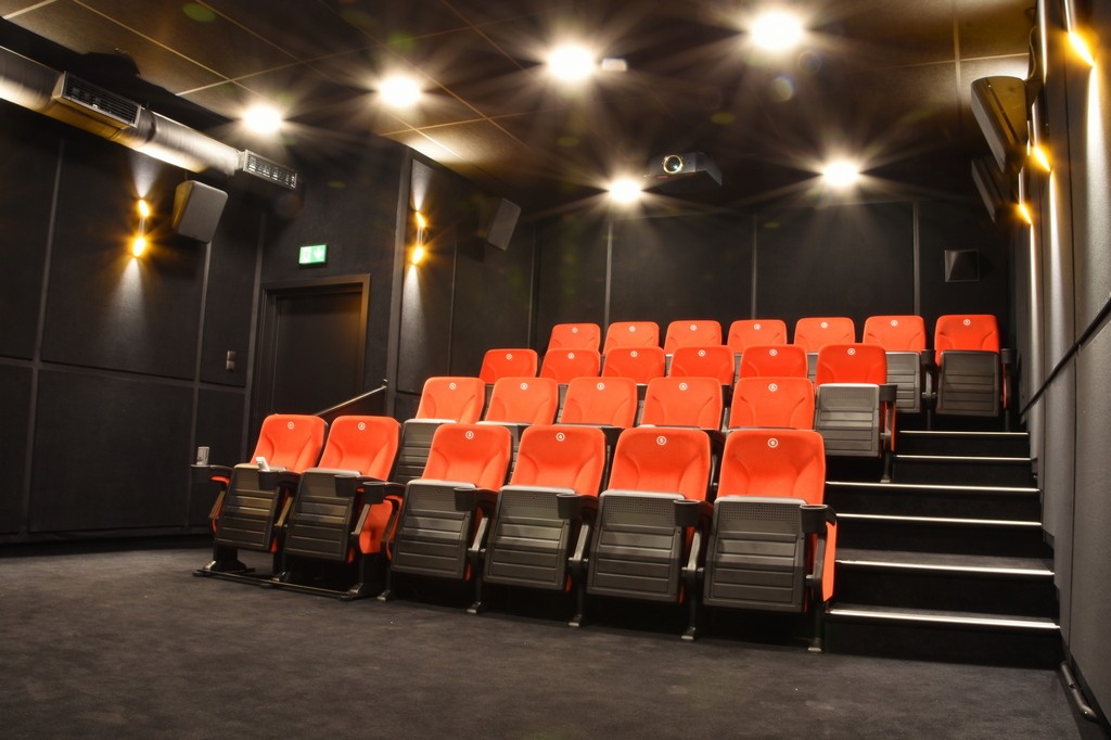 Małe, nowoczesne kino z rzędami czerwonych siedzeń, czarnymi podłokietnikami i przyćmionymi światłami sufitowymi. Kino jest puste, z tyłu znajduje się projektor, a dookoła czarne ściany.