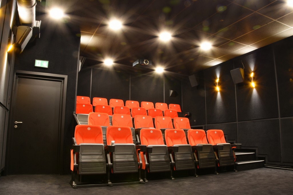 Małe nowoczesne kino z trzema rzędami pustych czerwonych siedzeń, czarnymi ścianami, delikatnymi światłami sufitowymi i projektorem zamontowanym na suficie. Po lewej stronie znajdują się drzwi wyjściowe.