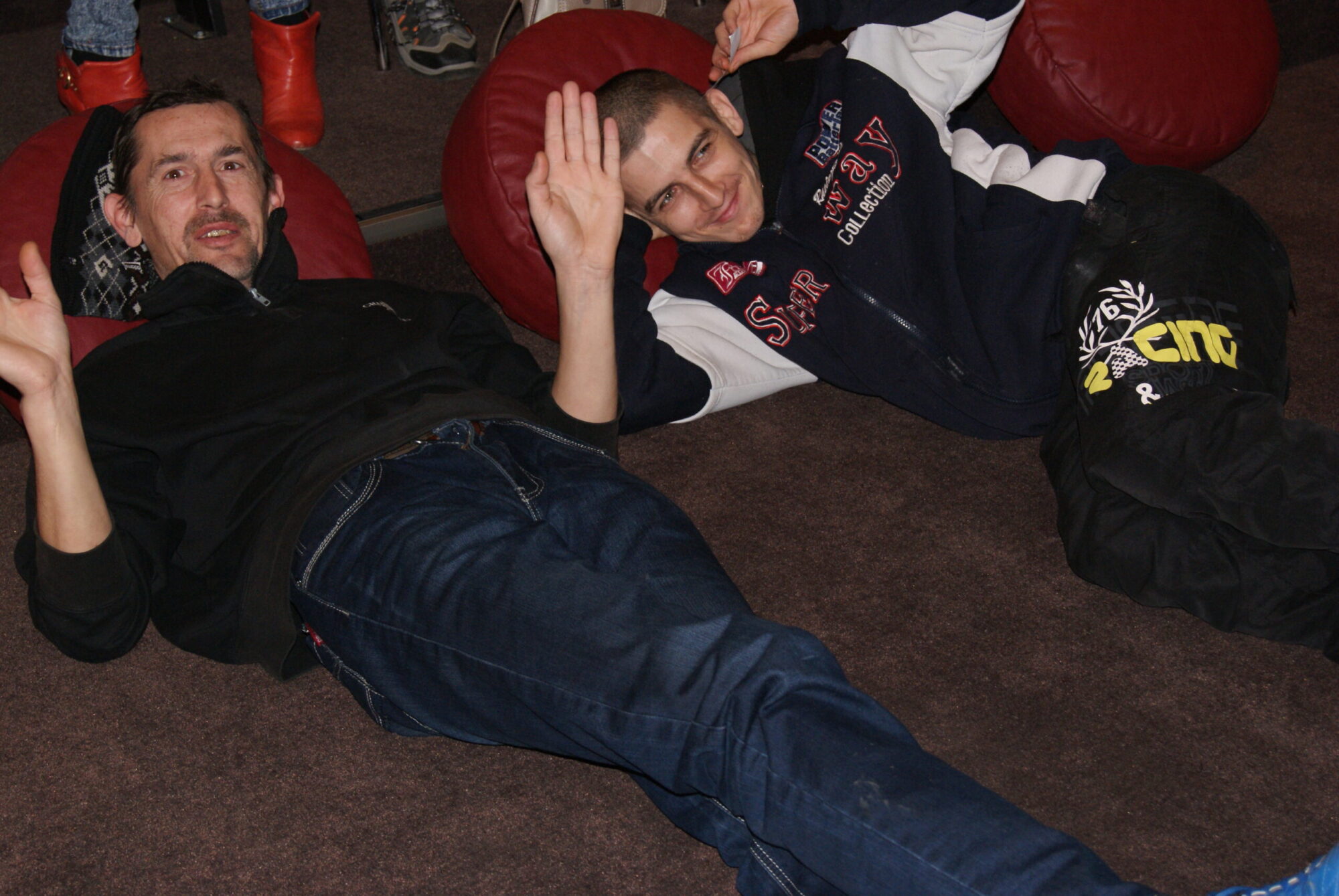 Dwóch mężczyzn leży na podłodze w pomieszczeniu, uśmiecha się i macha do kamery. Są zrelaksowani, obok czerwonych worków z fasolą i ubrani w swobodne ubrania. Otoczenie wydaje się wygodne i nieformalne.