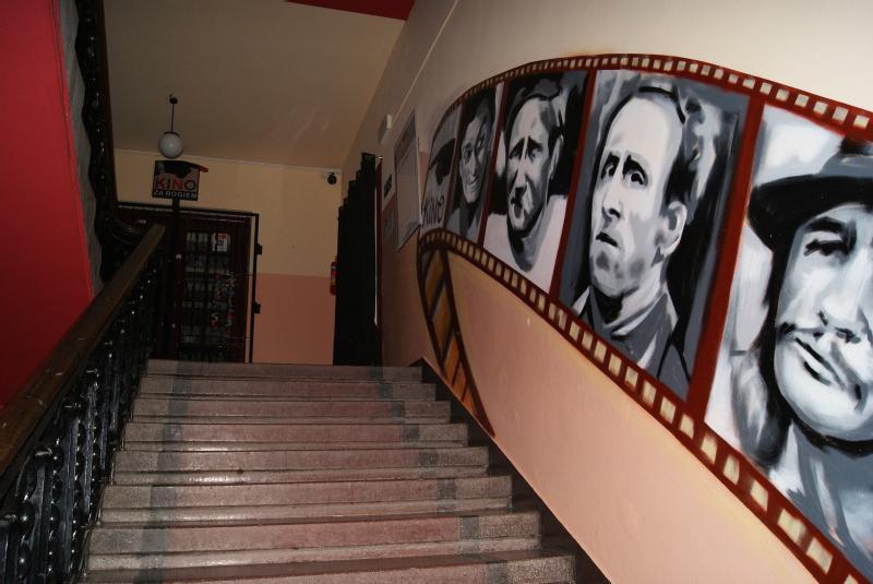 Schody z czarną poręczą prowadzą na górę; ściana obok nich jest ozdobiona dużymi, malowanymi portretami różnych osób w stylu filmstrip. Na szczycie schodów widoczny jest znak z napisem Kino.