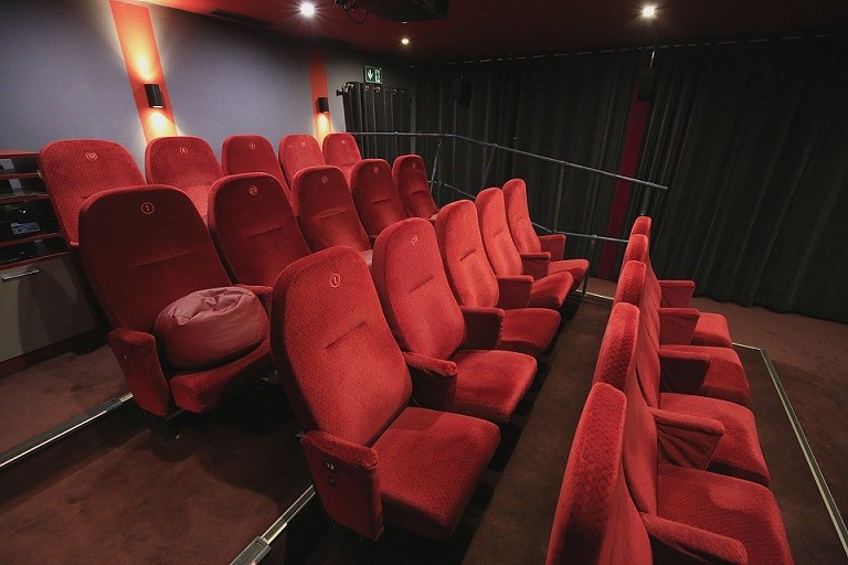 Rzędy pustych czerwonych foteli teatralnych są ustawione w małym, słabo oświetlonym kinie z ciemnymi ścianami i czarną zasłoną z przodu. Sala ma przytulne, intymne oświetlenie i nowoczesny design.