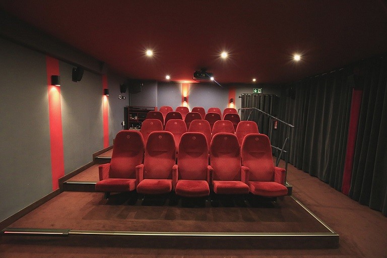 Małe, puste kino z czerwonymi, wyściełanymi siedzeniami ustawionymi w trzech rzędach, słabym oświetleniem, szarymi ścianami i projektorem widocznym na suficie.
