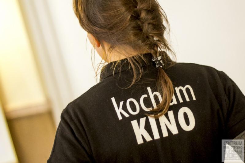 Osoba z warkoczami w kolorze brązowym jest widoczna od tyłu, ubrana w czarną koszulkę z białym napisem kocham KINO. Tło jest rozmyte i jasne.