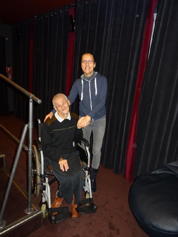 Starsza osoba na wózku inwalidzkim uśmiecha się, trzymając za rękę stojącą kobietę w okularach. Są w pomieszczeniu, przed ciemnymi zasłonami w pobliżu metalowej poręczy.