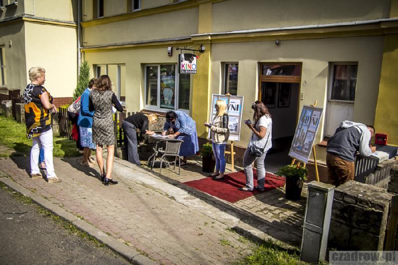 Mała grupa ludzi stoi i pochyla się nad stołem na zewnątrz budynku ze znakiem Kino, prawdopodobnie kupując bilety lub podpisując się. Plakaty filmowe są wystawione na sztalugach przy wejściu.