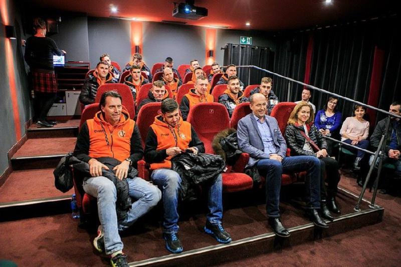 Grupa ludzi siedzi w małym teatrze z czerwonymi siedzeniami. Większość nosi pasujące do siebie pomarańczowe i czarne kurtki, podczas gdy kilka innych osób w pierwszym i ostatnim rzędzie nosi swobodne ubrania. Wygląda na to, że biorą udział w wydarzeniu lub prezentacji.