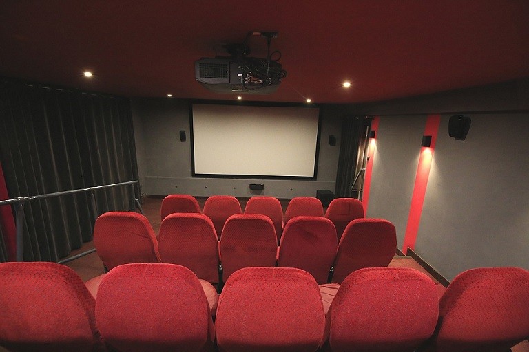 Małe kino z rzędami pustych czerwonych siedzeń zwróconych w stronę dużego, pustego ekranu, otoczone słabym oświetleniem i szarymi ścianami z czerwonymi akcentami. Nad siedzeniami widoczny jest projektor zamontowany na suficie.