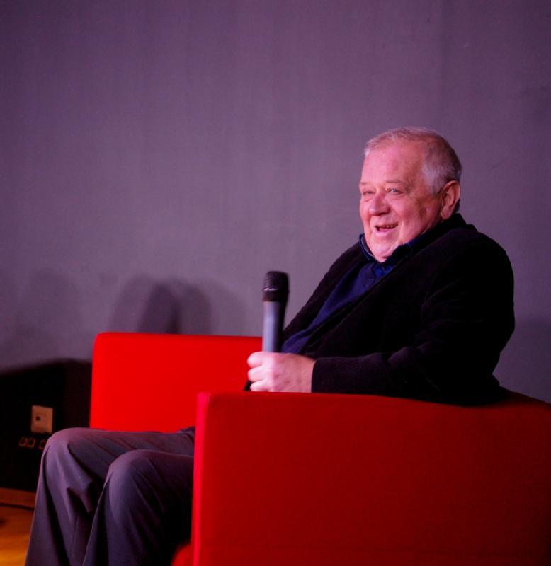 Starszy mężczyzna siedzi na jaskrawoczerwonym krześle, uśmiecha się i trzyma mikrofon, na jednolitym szarym tle.