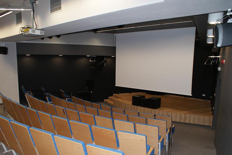 Pusta sala wykładowa z drewnianymi siedzeniami ustawionymi naprzeciwko sceny i dużym, białym ekranem projekcyjnym, ciemnymi ścianami i oświetleniem sufitowym.