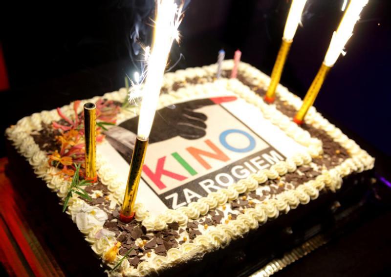 Prostokątny tort z białym lukrem i wiórkami czekoladowymi, z zimnymi ogniami i świecami. Na wierzchu tortu znajduje się nadrukowany jadalny obraz z napisem Kino Za Rogiem, kolorowymi literami i grafiką.