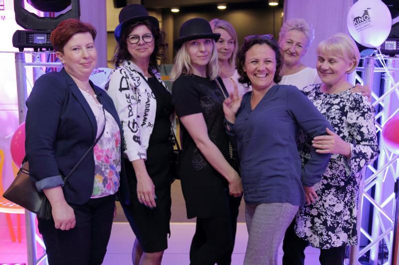 Grupa siedmiu kobiet pozuje razem w pomieszczeniu, niektóre uśmiechnięte, inne w kapeluszach, stojąc przed oświetloną ekspozycją z balonami i dekoracyjnymi światłami.