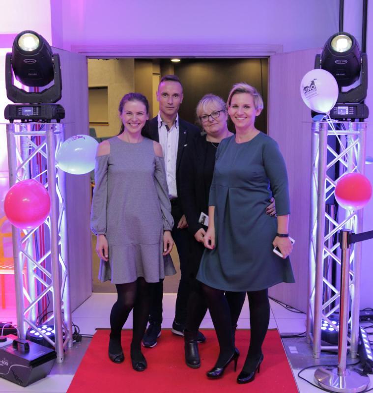 Cztery osoby pozują i uśmiechają się na czerwonym dywanie podczas imprezy w pomieszczeniu zamkniętym, otoczone balonami i światłami scenicznymi, przy fioletowo-różowym nastrojowym oświetleniu.